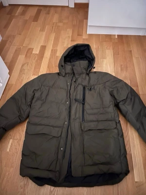 Olivgrön pufferjacka Helly Hansen XL - Olivgrön pufferjacka från Helly Hansen i storlek XL. Jackan har huva, stora fickor med lock, dragkedja och tryckknappar framtill samt HH-logga på bröstet och ryggen. Perfekt för kalla dagar med sin tjocka och isolerande design.