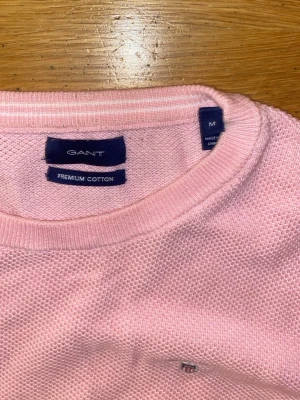 Rosa stickad tröja från GANT - Snygg ljusrosa stickad tröja från GANT i premium bomull. Tröjan har rund hals, lång ärm och en liten broderad logga på bröstet. Perfekt för dig som gillar klassisk och stilren design med en mjuk känsla.