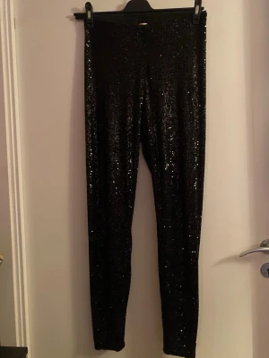 Svarta paljettleggings från H&M - Glittriga svarta leggings från H&M i storlek M, täckta med paljetter för extra bling. Stretchigt material som sitter tight och ger en snygg siluett. Perfekta när du vill sticka ut och glänsa lite extra. Supersnygga till sneakers eller boots. Dom är använda me slitet mellan benen
