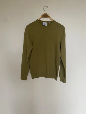  Stickad tröja  - Snygg olivgrön stickad tröja från Bläck med rund hals och ribbade muddar vid ärmslut och nederkant. 100% bomull 