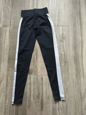 Svarta leggings med vita ränder Gina Tricot - Svarta leggings från Gina Tricot i storlek S med bred midja och vita ränder längs sidorna. Materialet är syntet och har en glansig finish med svarta ränder på ena benet. Perfekta för träning eller chill. Snygg, sportig look med tight passform.