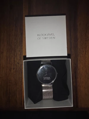 Silverfärgad meshklocka Klockjävel - Stilren klocka från Klockjävel of Sweden med svart urtavla och silverfärgat mesharmband i rostfritt stål. Minimalistisk design med rund boett och diskret logga på urtavlan. Perfekt accessoar för dig som gillar enkel och modern stil.