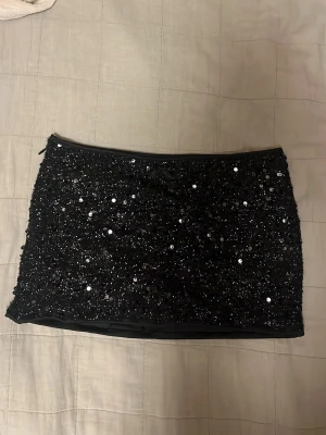 Svart paljettkjol med rosett - Säljer en svart kortkjol täckt med glittriga paljetter och en liten rosett upptill. Kjolen har en tight passform och inbyggda shorts 🥰Aldrig använt 