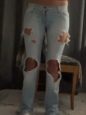 Ljusblå ripped jeans - Säljer ett par ljusblå jeans med stora ripped detaljer på både lår och knän. Jeansen har klassisk femficksdesign, knapp och dragkedja framtill samt raka ben. Perfekt för dig som gillar en edgy och avslappnad stil.