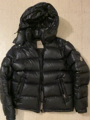 Svart Moncler pufferjacka med huva - Hej! Jag säljer nu min Moncler Maya då den har blivit för liten för mig.Storlek 3 motsvarar M/L,jackan är som ny, inga defekter alls (hål,slitage). Jackan är köpt på NK för 15.999kr. Hör gärna av er vid eventuella frågor!