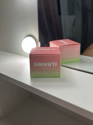 Smuuti Skin Watermelon Jelly Cream - En söt ansiktskräm med gelétextur från Smuuti Skin, Watermelon Jelly Cream. Kommer i en rund burk med rosa lock och grön-rosa ombre design. Innehåller vattenmelonextrakt och hyaluronsyra för återfuktning. Perfekt för dig som gillar fräscha och fruktiga hudvårdsprodukter.