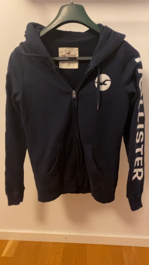 Blå hoodie från Hollister med tryck - Svart hoodie från Hollister med dragkedja, huva och stora vita bokstäver längs ena ärmen. Klassisk logga på bröstet och tryckt detalj på huvan. Perfekt för en chill och avslappnad stil.