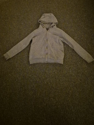 Grå mckenzie zip tröja  - Säljer en grå hoodie från McKenzie med huva och hel dragkedja framtill. Tröjan har långa ärmar, ribbade muddar och tryckt logga på bröstet. Perfekt för chill eller träning. Materialet är mjuk bomullsmix som känns skönt mot huden.