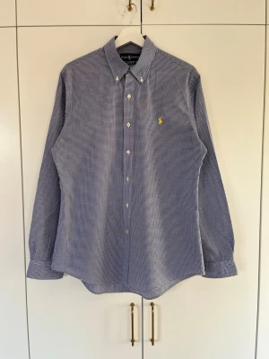 Ralph Lauren skjorta  - Elegant Ralph Lauren skjorta i blått och vitt med Custom Fit passform. Button down krage och ikonisk broderad logga i gult. Ett säkert och tidlöst val för en stilren look. Hör gärna av dig vid frågor eller funderingar 😄