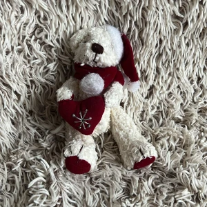 Jul nallebjörn - En söt nallebjörn med tomteluva, en röd halsduk och björnen håller i ett rött hjärta med broderad snöflinga. Nallen är vit med röda detaljer 