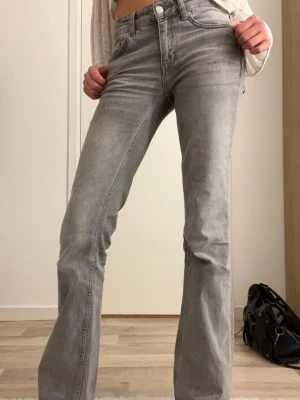 Grå bootcut jeans med hög midja - Snygga grå jeans med bootcut-snitt och hög midja. Jeansen har klassisk femficksdesign, bälteshällor och knappstängning fram. Materialet är mjukt denim i en ljusgrå ton som ger en cool vibe. Perfekta för dig som gillar en retrostil med modern touch.