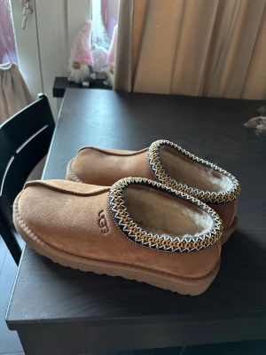 UGG slip-on mockasiner med mönstrad kant - Bruna slip-on mockasiner från UGG med mjuk fårullsfoder och grov platt sula. Skorna har en snygg kant med gul, vit och brun zigzag-mönster runt öppningen. Perfekt för chill dagar och mysiga stunder hemma. Storlek 40, men skulle passa någon med storlek 39 också. 