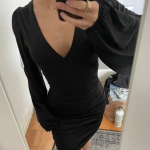 Svart klänning med v-ringning - Snygg svart klänning med v-ringning och långa ärmar. Den har en tight passform och rynkade detaljer längs sidorna. Super bekväm och endast ANVÄND 1 GÅNG, nästintill nyskick!☺️