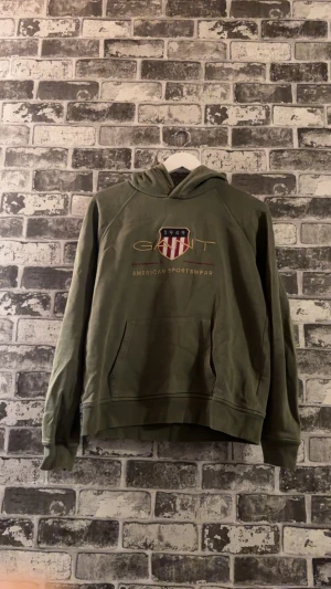 Gant hoodie - Tröjan är en olivgrön GANT-hoodie. Den har en logotyp i guld som består av ordet "GANT" ovanför en sköld med röda och vita ränder, årtalet "1949" och texten "AMERICAN SPORTSWEAR" broderat under skölden.