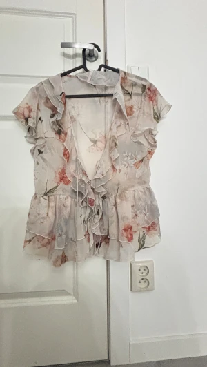 Blommig volangblus från Zara - Blus från Zara i storlek S. Blusen är ljusbeige med blommigt mönster i rosa och grönt, har volanger längs halsringningen och axlarna samt snörning framtill. Materialet är tunt och luftigt.
