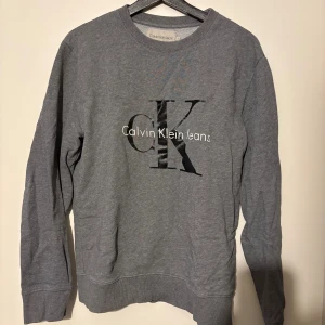 Grå sweatshirt från Calvin Klein Jeans - Säljer en stilren grå sweatshirt från Calvin Klein Jeans i storlek S. Tröjan har ett stort CK-tryck på bröstet och rund halsringning med ribbade muddar vid ärmslut och nederkant. Perfekt för en chill och avslappnad look.