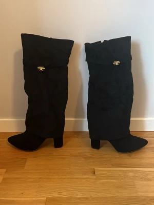 Svarta mocka stövlar med spetsig tå - Säljer ett par svarta mocka stövlar med spetsig tå och bred klack. Stövlarna har guldfärgade detaljer och dragkedja på insidan. Materialet är mocka och de har en stilren look som passar till många outfits. So storleken är 40 men de behövs lägga in en sula så då blir storleken 39. Min syster som har 39 passade henne då. Detta var ett felköp för mig med storlek så jag vill endast få de sålda