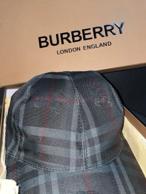 Rutig Burberry keps i bomull - Säljer en snygg Burberry keps i klassiskt rutigt mönster med inslag av grått, svart och röda linjer. Kepsen är tillverkad i bomull och har justerbar rem baktill. Perfekt accessoar för dig som vill ha en stilren och lyxig look.