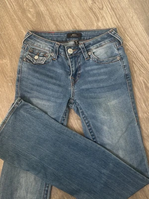 True Religion Becca Bootcut Jeans blå - Säljer ett par blå jeans från True Religion, modell Becca Mid Rise Bootcut. Jeansen har klassisk femficksdesign, snygga kontrastsömmar och detaljerad bakficka med logga. Materialet är slitstarkt denim i bomull och passformen är bootcut med midwaist.