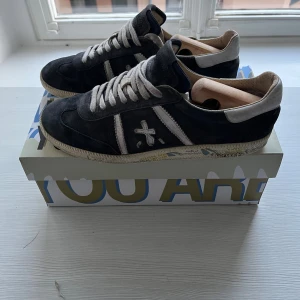 Premiata blå sneakers i mocka - Blåa sneakers från Premiata med beige detaljer och snörning. Skorna har en rund tå, platt sula och är tillverkade i mjuk mocka med kontrasterande vita partier och logotyp på sidan och hälen. Kommer med låda.