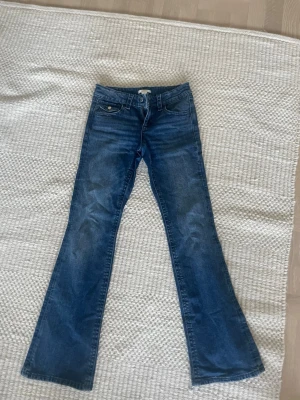 Blå bootcut jeans med rosett-detalj - Helt nya snygga blå jeans med bootcut-snitt och coola broderade rosetter på bakfickorna. Klassisk femficksmodell med knapp och dragkedja framtill. Jeansen har en mörk tvätt och är tillverkade i jeansmaterial som ger en skön passform.