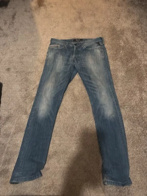 Replay jeans i blå tvätt, storlek 30 - Säljer ett par snygga Replay jeans i klassisk blå tvätt med slitningar på låren och knäna. Jeansen har rak passform och normal midja, med Replay-logga på insidan. Materialet är robust jeans och de har klassiska fem fickor och knappgylf. Ett igensytt hål i skrevet men det tänker man inte på när man ser dom