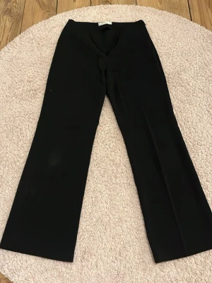 Svarta wide kostymbyxor från Friendtex - Säljer ett par svarta kostymbyxor från Friendtex i storlek 38. Byxorna har en wide passform med raka ben och är tillverkade i ett slätt material som ger en stilren look. Perfekta för dig som gillar klassisk och enkel stil. Det är även små, små ränder på byxorna.