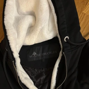 Svart teddyjacka Moose Knuckles - Svart teddyjacka från Moose Knuckles med vit fluffig fuskpäls på insidan av huvan. Jackan har dragkedja, snörning i huvan och ett snyggt logomärke på ärmen. Insidan är quiltad med mönstrat foder. Perfekt för kalla dagar. Är äkta. Har tag i jackan och har legit checkat den👍