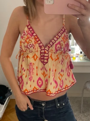 Beige och rosa mönstrad linnetopp - Säljer en beige linnetopp med färgstarkt mönster i rosa och orange. Toppen har smala axelband, v-ringning med dekorativa detaljer och pärlor. Jag har knutit axelbanden där bak för att den ska passa mig men de går såklart att ändra!