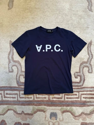  A.P.C. - Snygg mörkblå t-shirt från A.P.C. med stort vitt tryck på bröstet. Klassisk rund hals och korta ärmar. Tillverkad i mjuk bomull som känns skön mot huden. Perfekt för dig som gillar stilrena och enkla plagg med tydlig logga.
