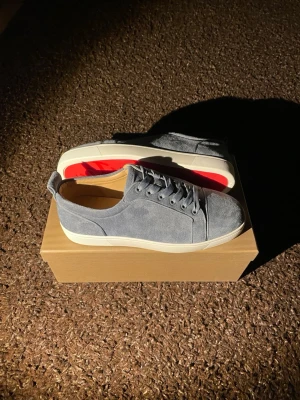 Grå mocka sneakers Christian Louboutin - Säljer ett par gråa sneakers från Christian Louboutin i mjuk mocka med klassisk röd sula och vita kanter. Skorna har rund tå, snörning och beige innersida. Kommer med originalpåse och certifikat från Paris.
