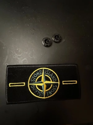 Stone Island svart västpatch - Säljer en klassisk svart Stone Island patch med broderad kompasslogga i gult och grönt. Två svarta knappar med Stone Island-tryck ingår. Perfekt att fästa på din väst för att ge den en ikonisk streetwear-look.