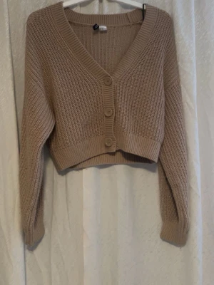 Beige croppad stickad kofta - Säljer en beige croppad stickad kofta med v-ringning och stora knappar framtill. Koftan har långärmade ribbade muddar och ett grovt stickat mönster som ger en chill vibe. Perfekt att slänga över en t-shirt eller linne för extra lager.