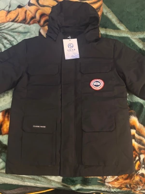 Svart parkajacka med huva och päls - Svart parkajacka från Classic Vogue med stor huva och avtagbar pälsdetalj. Jackan har fyra rejäla fickor framtill, dragkedja och tryckknappar. Märke med röd och vit logga på bröstet. Perfekt för kalla dagar och har en clean, urban look.          Jag har köpt denna jacka men den var för liten, så jag tänker sälja den.