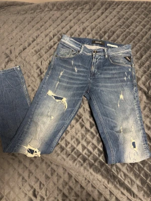 Replay Broken Edge blå jeans - Säljer ett par Replay Broken Edge jeans i klassisk blå tvätt med slitna detaljer och hål på benen. Modellen är slim tapered fem fickor och coola slitningar både fram och bak. Materialet är jeans och passformen är normal. Perfekt för dig som gillar en edgy streetstil.
