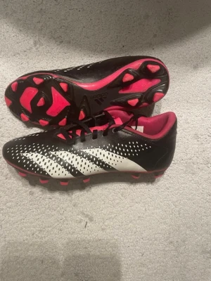 Adidas fotbollsskor svart/rosa strl 40 - Säljer ett par Adidas fotbollsskor i storlek 40 med svart och vit ovandel, rosa detaljer och dobbar. Skorna har snörning och är tillverkade i syntetmaterial. Insidan är fodrad i rosa och yttersulan har mönstrade dobbar för bra grepp på planen.