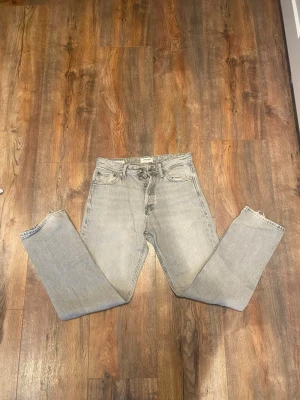 Grå relaxed jeans från Jack & Jones - Snygga ljusgrå jeans från Jack & Jones i modellen Relaxed/Chris. Jeansen har en avslappnad passform, raka ben och klassisk femficksdesign. Tillverkade i mjukt bomullsmaterial med en tvättad look och metallknappar. Perfekta för en chill och stilren vibe.