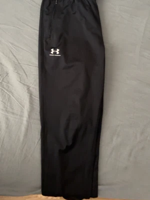 Svarta träningsbyxor från Under Armour - Säljer ett par svarta träningsbyxor från Under Armour med dragsko i midjan och dragkedjor vid fickorna. Byxorna har en stilren look med logga på vänster ben och är gjorda i ett mjukt syntetmaterial som är perfekt för sport och chill.