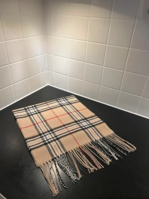 Beige rutig halsduk - Säljer en beige halsduk med klassiskt rutigt mönster i svart, vitt och rött. Halsduken har tunna fransar i ändarna och är mjuk och skön att ha runt halsen. Perfekt för att ge din outfit en snygg touch.