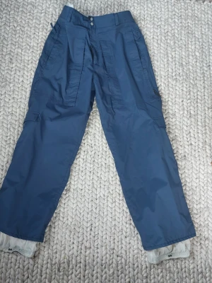 Blå täckbyxor från Burton Ronin L - Säljer ett par blå täckbyxor från Burton Ronin i storlek L. Byxorna har flera fickor, elastiska benslut. Det finns en lagning på byxan vid fickan fram se bild.
