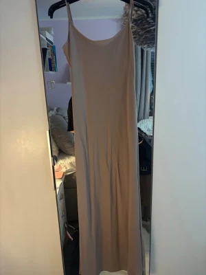 Beige långklänning med smala axelband - En beige långklänning från Bershka med smala axelband och rundad halsringning. Klänningen har en rak och enkel siluett.