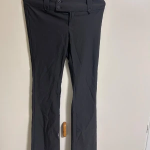 Gråa kostymbyxor med knappar - Snygga gråa kostymbyxor med låg/mellan midja och raka ben. Byxorna har flera dekorativa knappar i midjan och bakfickor med lock. Materialet känns mjukt och har en lätt stretch, perfekt för en clean och stilren look.