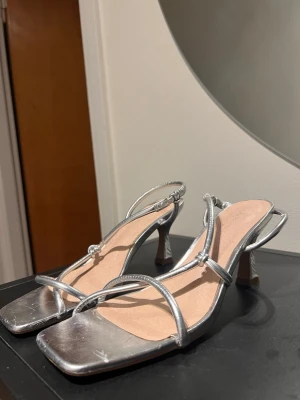 Silvriga sandaler med klack - Säljer ett par stilrena sandaler från asos i metallic silver. Skorna har smala remmar, fyrkantig tå och en låg klack. 