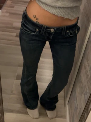 True Religion Joey Low Rise Flare Jeans - Säljer ett par blå True Religion Joey jeans med låg midja och flare-ben. Klassiska kontrastsömmar, snygga fickdetaljer med stora knappar och ikonisk patch bak.