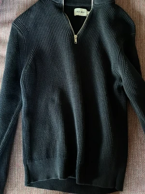 Svart stickad half zip från East West - Säljer en svart stickad tröja från East West med half zip-dragkedja och hög krage. Tröjan har ribbad struktur och långa ärmar, perfekt för kyligare dagar. Enkel och stilren design som funkar till det mesta.