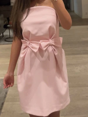 Ljusrosa axelbandslös kortklänning - Superfin ljusrosa axelbandslös klänning med två stora rosetter i midjan. Klänningen är kort och har en stilren, lite voluminös form. Perfekt för dig som vill ha en söt och trendig look. Endast använd i ett par timmar, precis som ny 💕