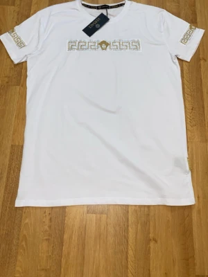 Vit  t-shirt med guldmönster - Vit t-shirt  med korta ärmar och exklusiva detaljer i guld och silver. Framsidan har ett broderat Medusa-motiv och geometriskt mönster med paljetter, som även återfinns på ärmarna. Tillverkad i mjuk bomull och har märke -logga i guld vid nederkanten.