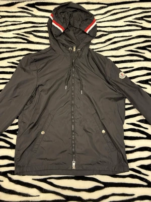 Moncler windbreaker - Svart  windbreaker från Moncler med huva som har röda, vita och mörkblå ränder. Jackan har dragkedja framtill, snörning vid huvan och två fickor med tryckknappar. Moncler-logga på vänster ärm och innerficka med märkning. Perfekt för dig som gillar stilren design. NFC Scan funkar bild  4 + påse och ett kort följes med , hör av dig vid minsta fundering 😌