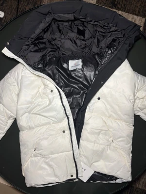 Vit Moncler pufferjacka med svart huva - Säljer en clean pufferjacka från Moncler i vit med svart huva och insida. Jackan har stora fickor, dragkedja och tryckknappar framtill. Moncler-logga broderad på huvan. Perfekt för dig som vill ha en stilren vinterlook.