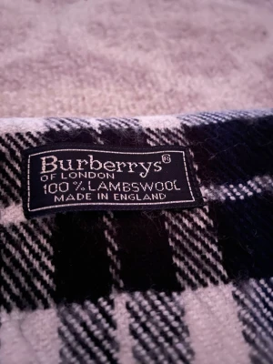 Rutig blå halsduk från Burberrys - Säljer en klassisk rutig halsduk från Burberrys of London, tillverkad i 100% lammull. Halsduken har en snygg kombination av marinblått, vitt, svart och en diskret röd linje. Materialet är mjukt och värmande, perfekt för kyliga dagar. Stilsäker design med fransar längs nederkanten.